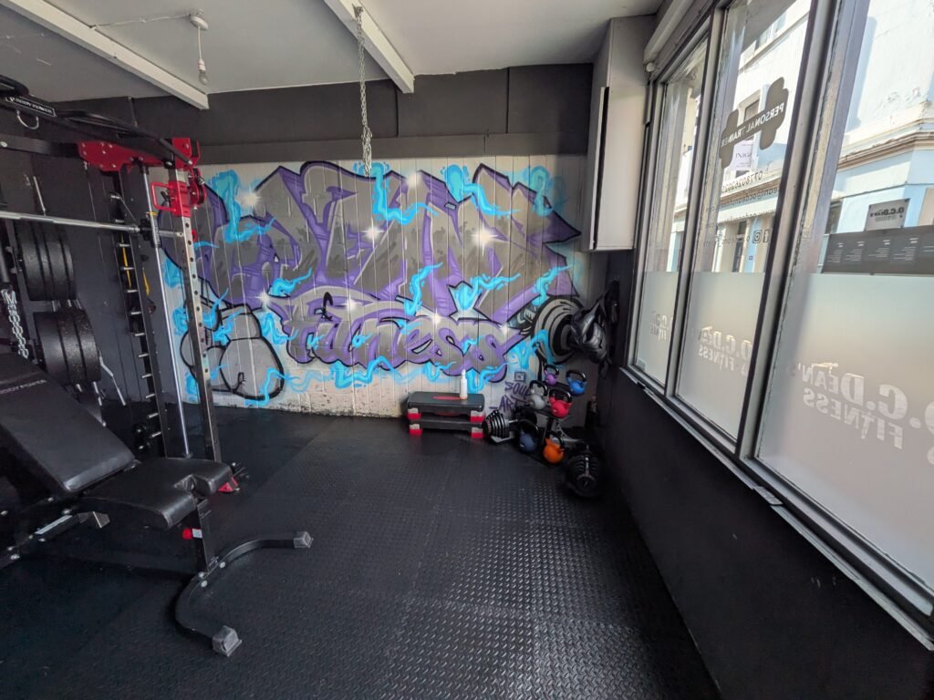 Interior of O.C. Dean's Fitness Margate, private gym with power rack, bench, kettlebells and a large purple and blue graffiti wall